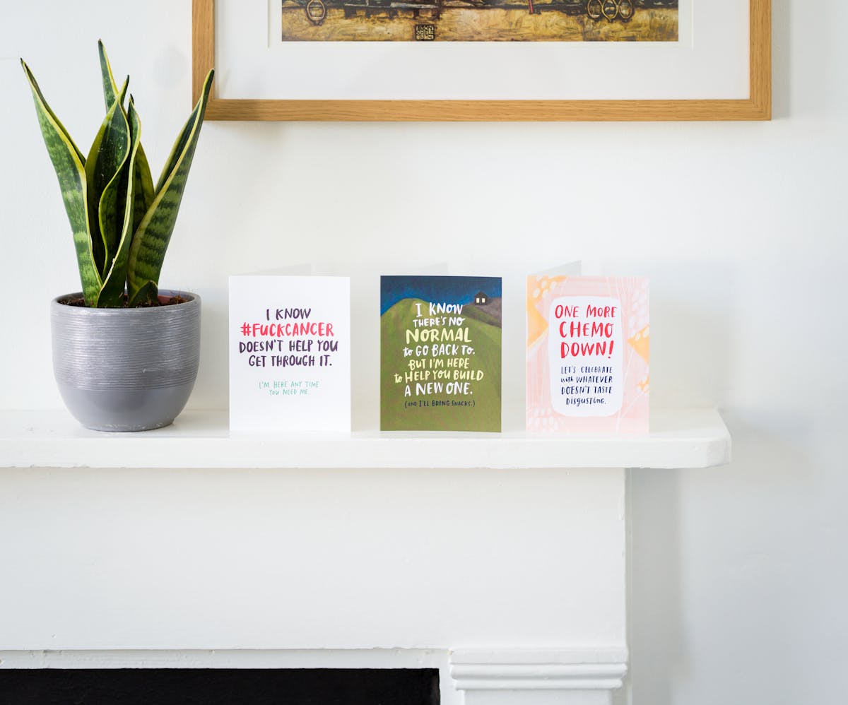 Photograph of a white mantlepiece against a white wall. On the mantlepiece is a cactus plant in a silver plant pot, next to which there are three greetings cards. One card reads, 'I know #fuckcancer doesn't help you get through it, I'm here anytime you need help'. The next reads, 'I know there's no normal to go back to but I'm here to help you build a new one (and I'll bring snacks)'. The last reads, 'One more Chemo down! Lets celebrate with whatever doesn't taste disgusting'.