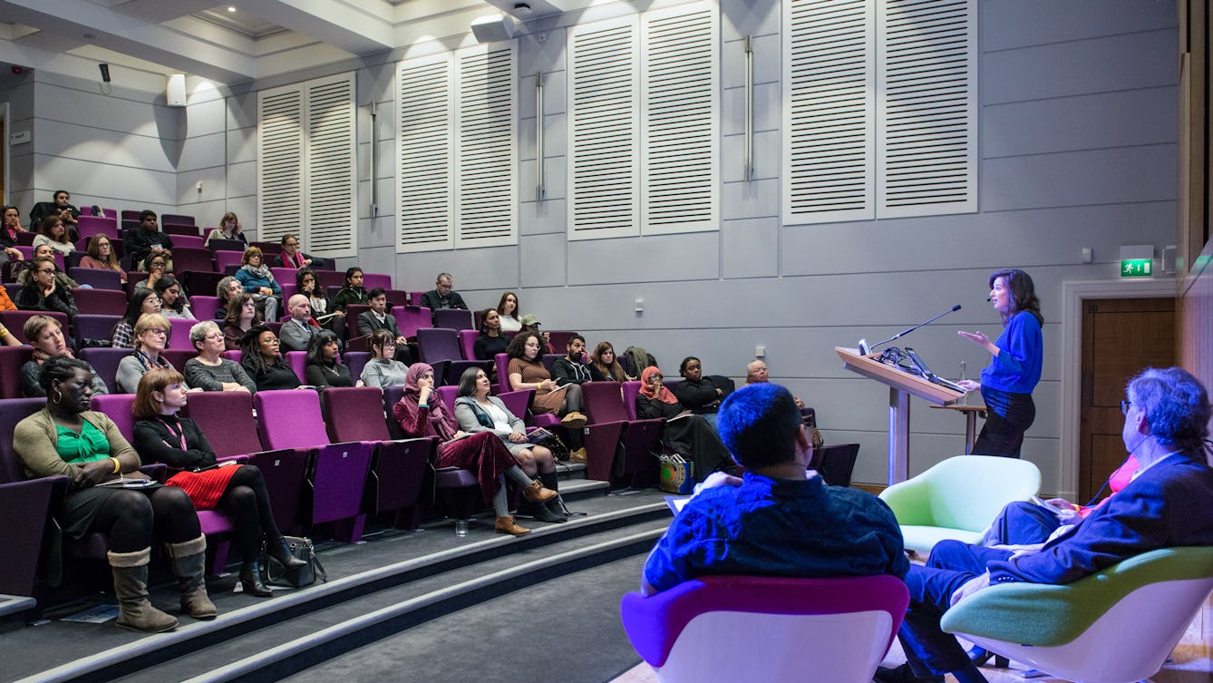 Colour photograph of people sitting in the Henry Wellcome Auditorium at Wellcome Collection on purple flip-down seats. They look towards a woman standing at a podium speaking and gesturing.