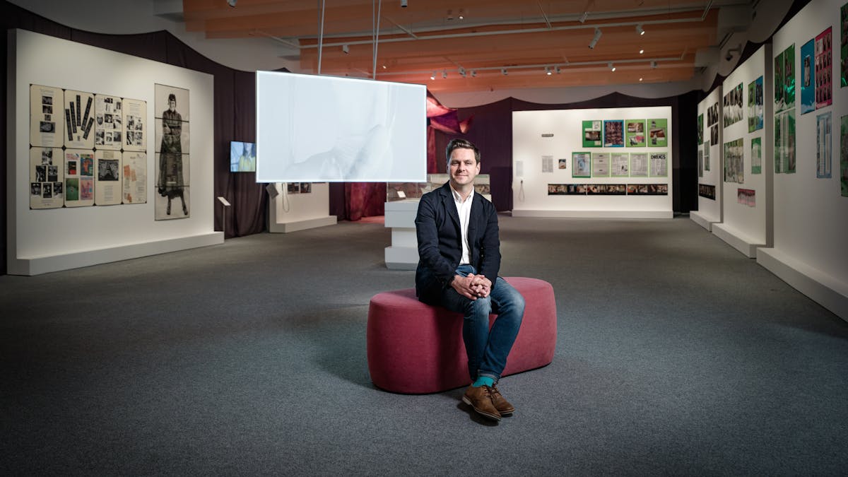 Photograph of Misbehaving Bodies curator George Vasey sitting in the exhibition space. Behind him are the works of Jo Spence; in the form of photographs and tear sheets hung on white panels, and Oreet Ashery; in the form of a projection of a white figure moving against a white screen.