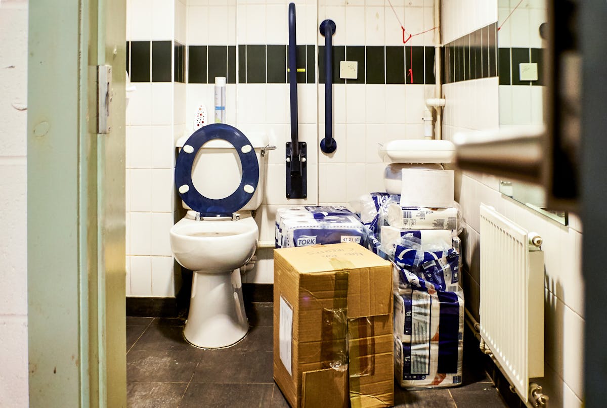 Photograph though a doorway into an accessible toilet. The floorspace next to the toilet is covered with a cardboard box and piles of packets of toilet paper.