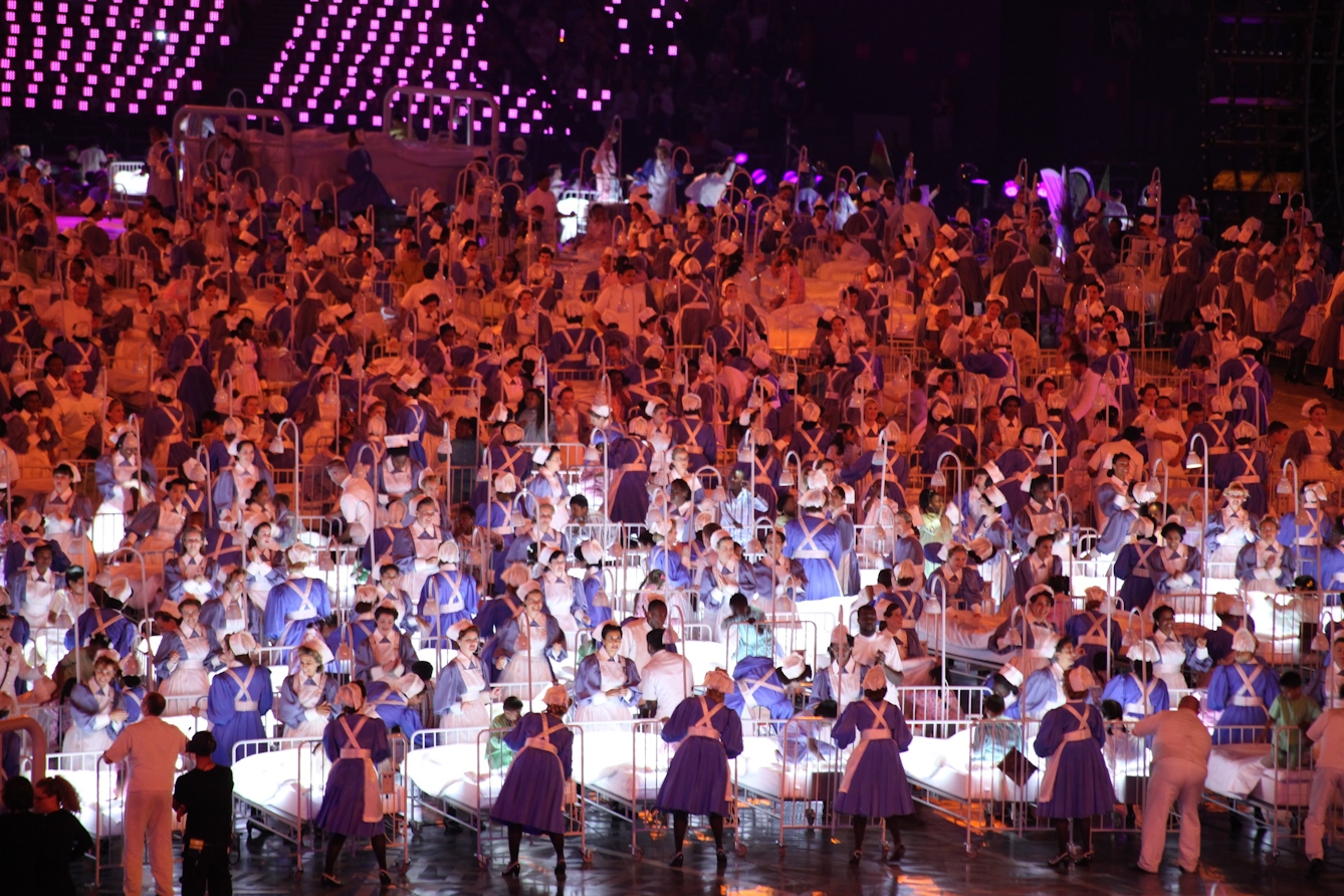 Photograph of many people dressed in historic nursing uniform of blue dresses and bonnets wheeling hospital beds containing children around as part of the 2012 Olympics Opening Ceremony.