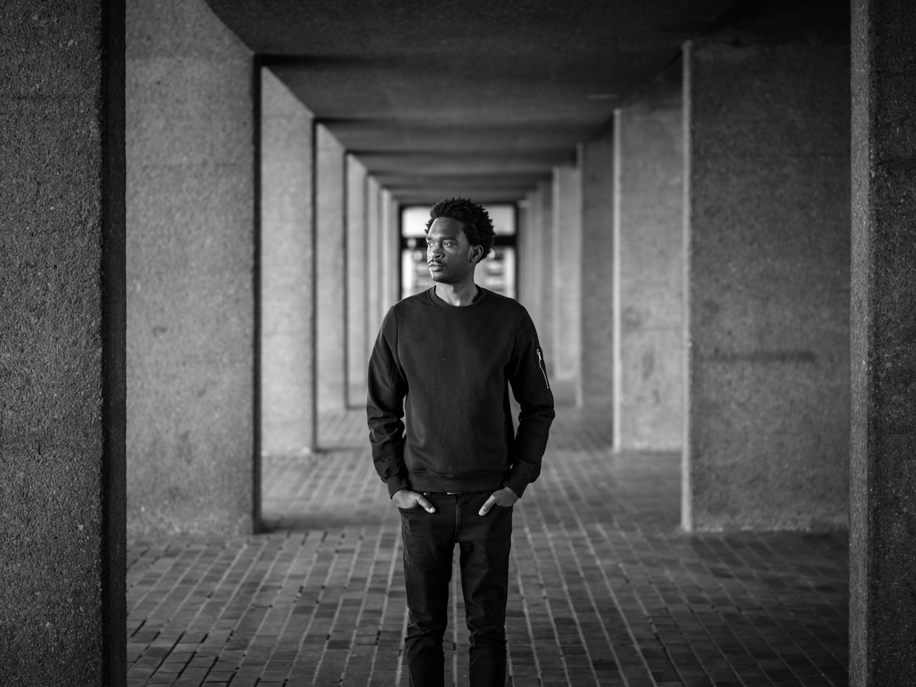 Photograph in black and white of a man standing in a walkway, looking out at the Barbican Centre, London.