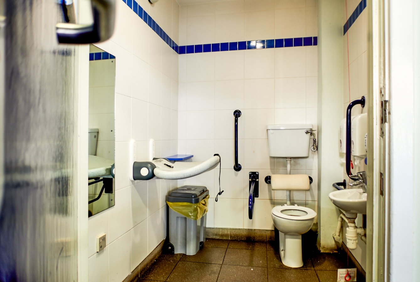 Photograph though the doorway of a run down accessible toilet showing part of the door and doorframe, along with the contents of the room, toilet, basin, sanitary bin and baby changing table.