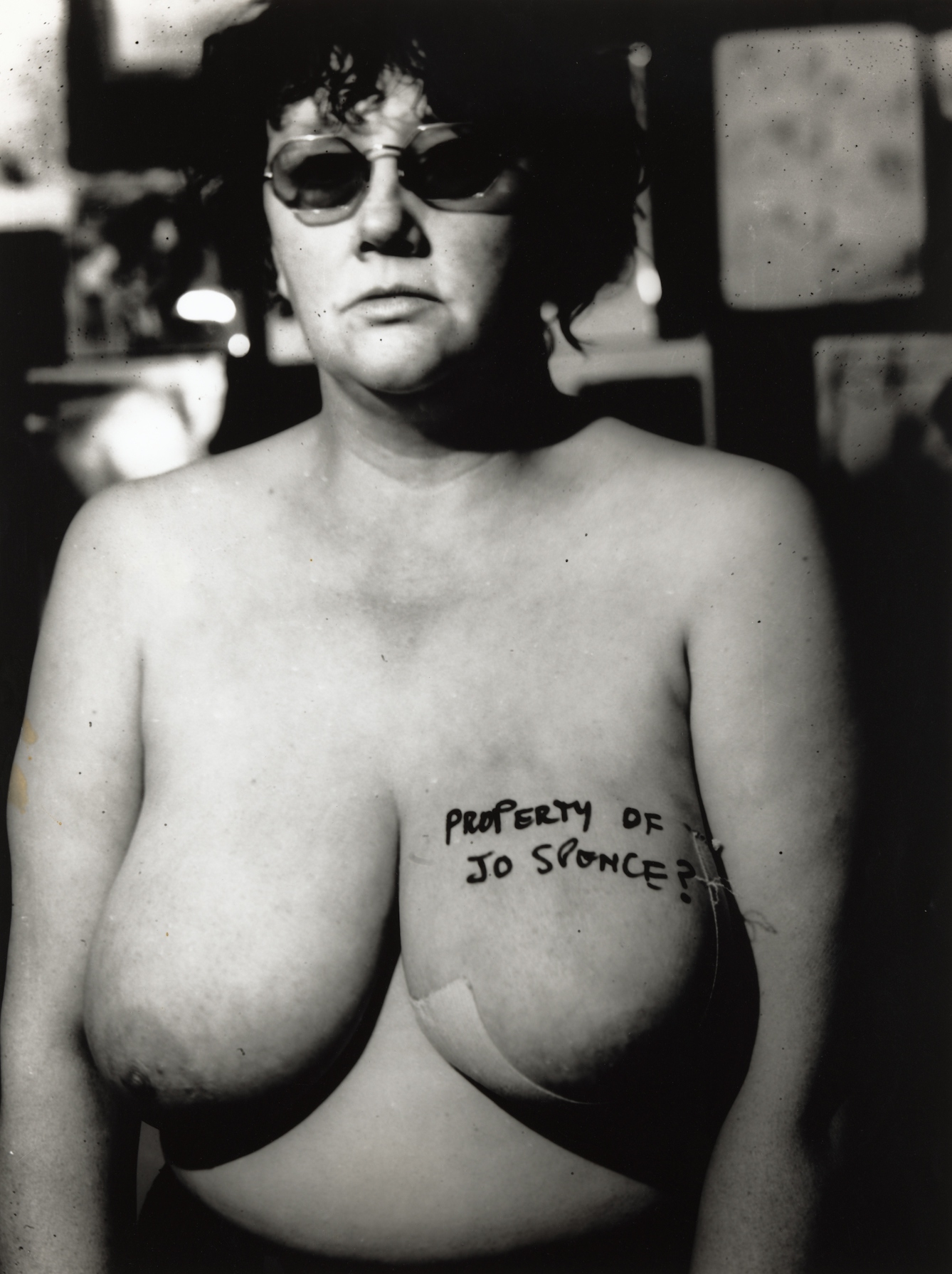 Black and white photograph of a woman wearing tinted glasses, naked from the waist up. on her left breast is written in black pen, "Property of Jo Spence?"
