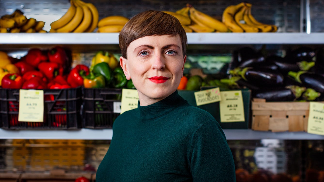 Photographic portrait of Elsa Richardson in front of a display of fruit and vegetables in a whole food shop.
