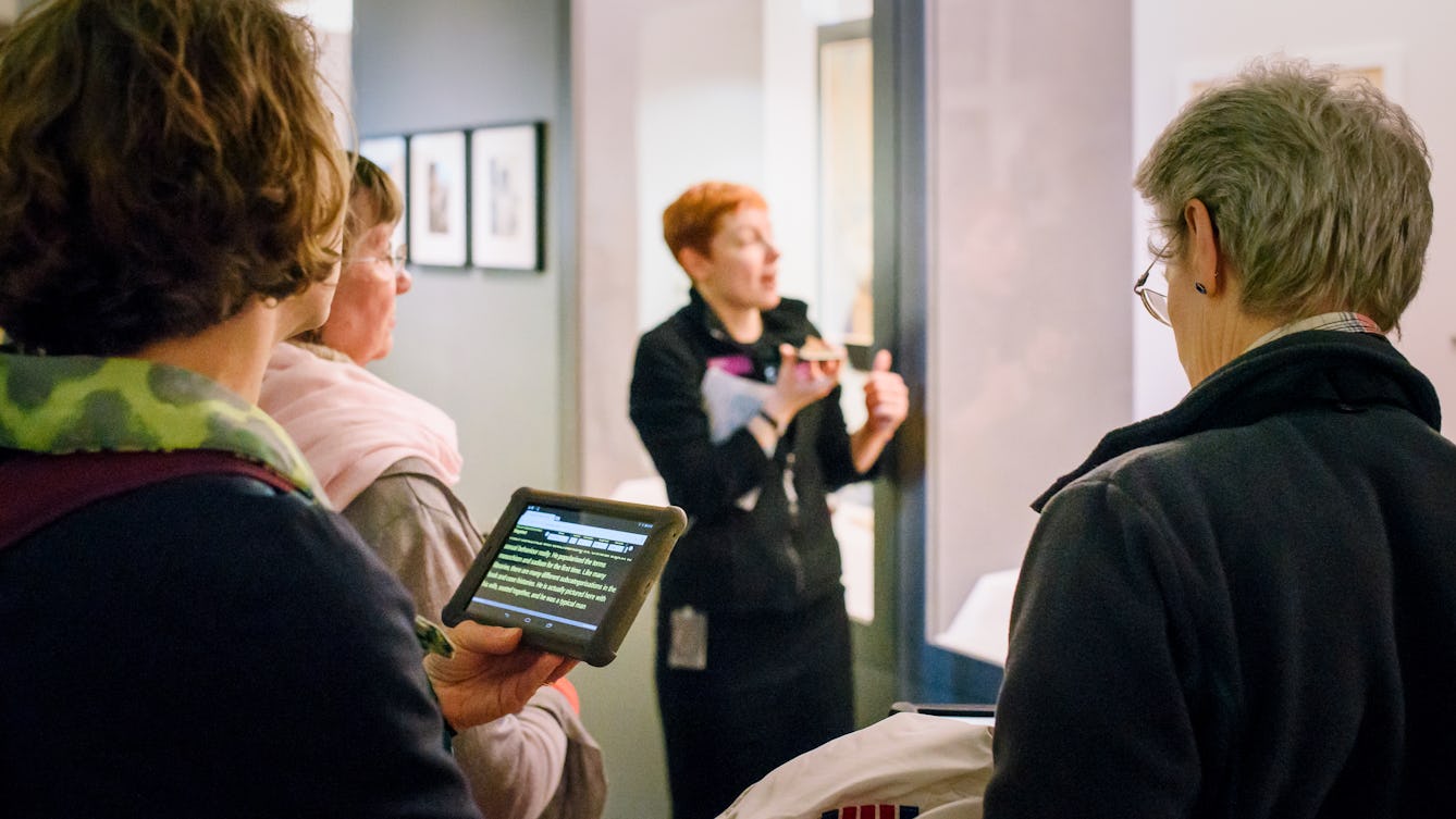 Photograph of a Wellcome Visitor Experience Assistant speaking into a phone whilst giving a speech-to-text tour of a gallery. In the foreground is a tablet displaying the transition.