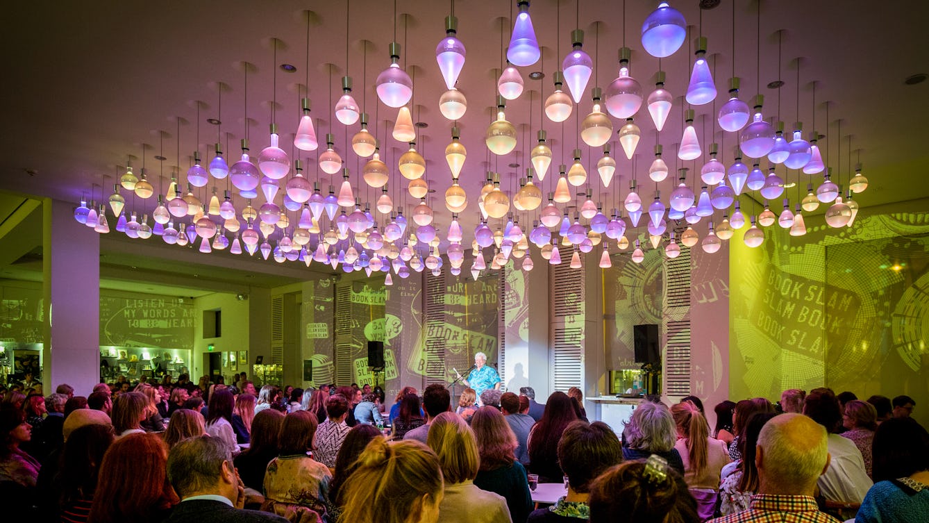 Photograph showing an audience listening to a spotlit speaker in the Wellcome Café, under colourful ceiling lights.