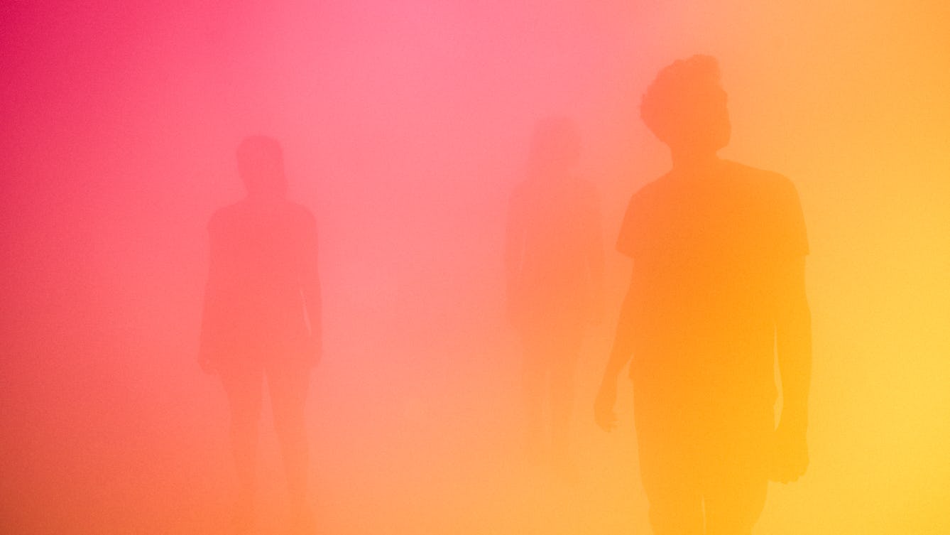 Photograph of three silhouettes in a room full of mist, bathed in a yellow and pink light.