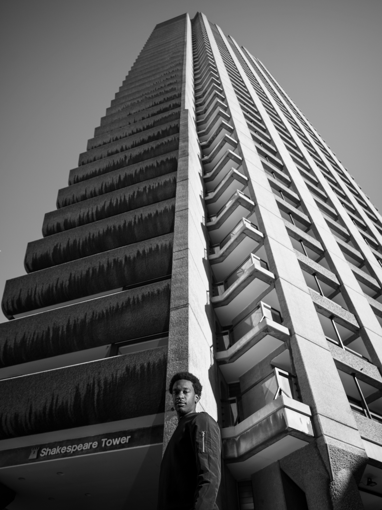 Photograph in black and white of a man looking down to camera with a high tower block soaring into the sky behind him at the Barbican Centre, London.