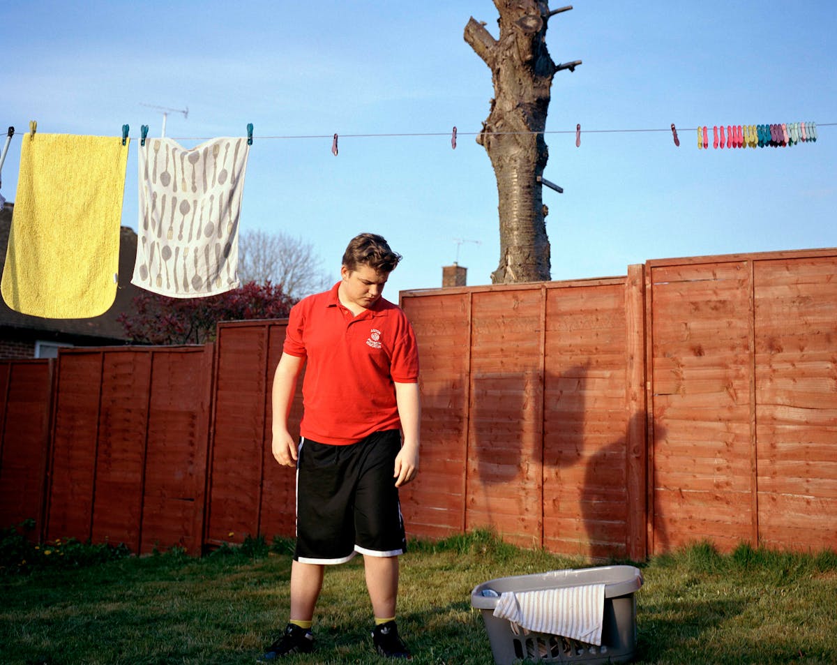 Photograph of a teenage boy wearing a red t-shirt and black shorts, standing in a garden. Above him is a washing line with pegs and towels.
