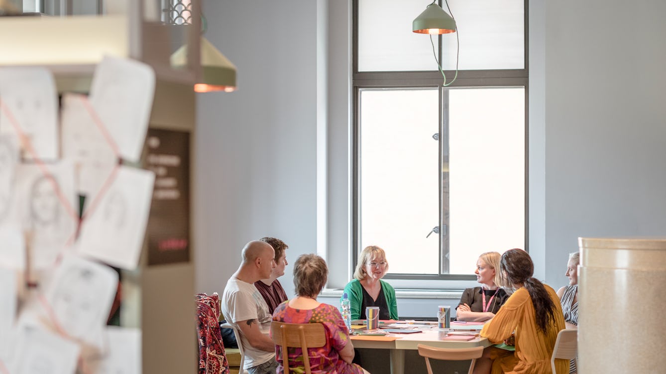 Photograph of a group of seven people sitting around a table in the Reading Room in Wellcome Collection. They are in discussion. Behind them is a large window and in the foreground is part of one of the Reading Room display cases.