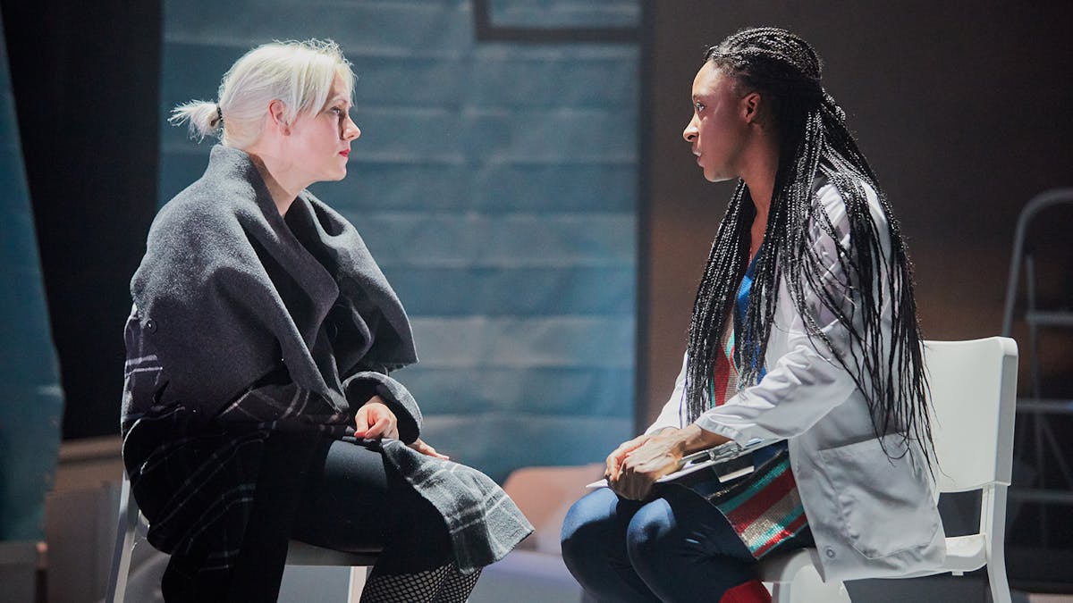 Two women sit opposite each other. One woman wears a lab coat and holds a clipboard, the other is wrapped in a large shawl.