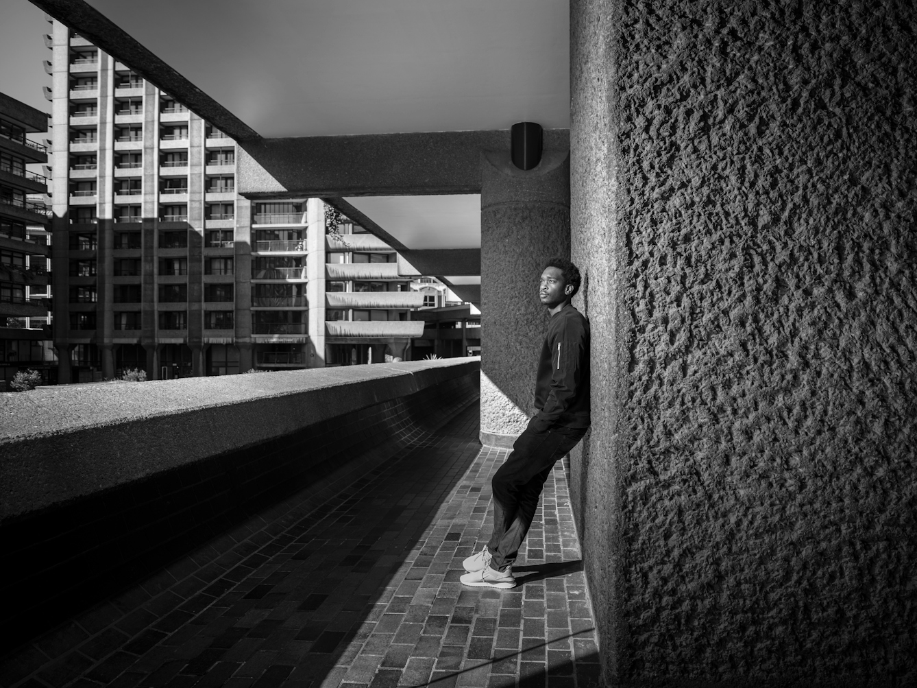 Photograph in black and white of a man standing in a walkway, looking out at the Barbican Centre, London.