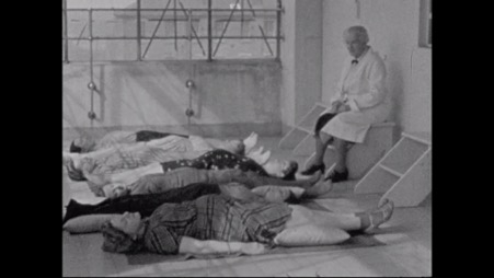 Image of women lying on their backs in a line. A man wearing a doctor's coat sits nearby, looking at them.