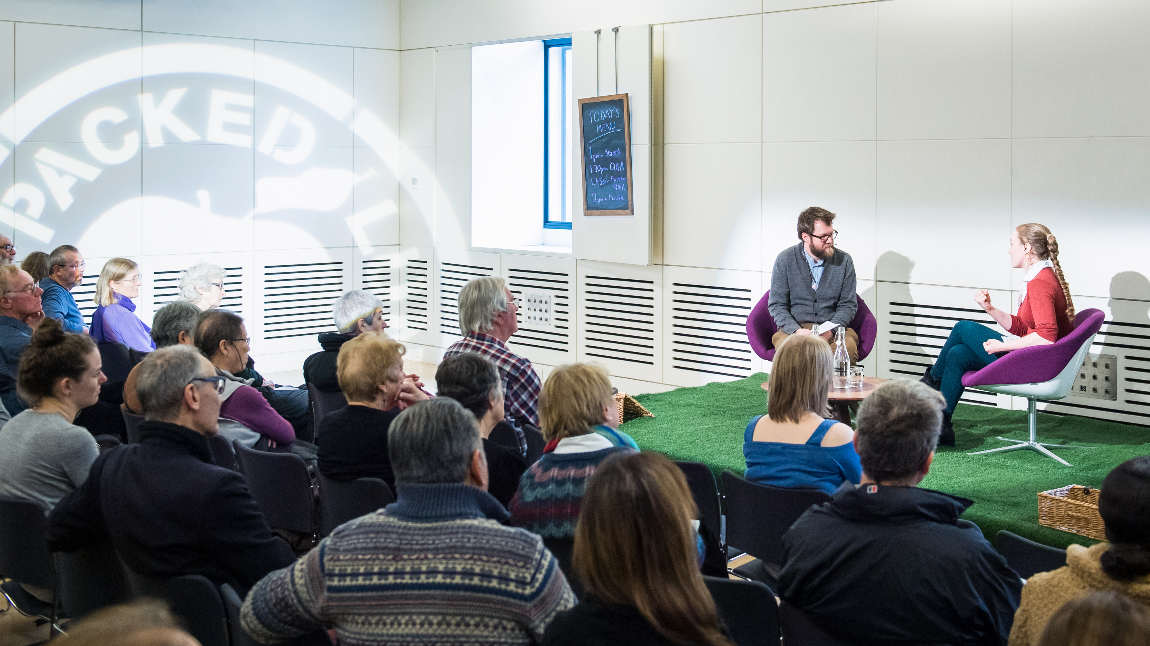Photograph of a man and a woman sitting on a stage, in conversation. In front of them is an audience.
