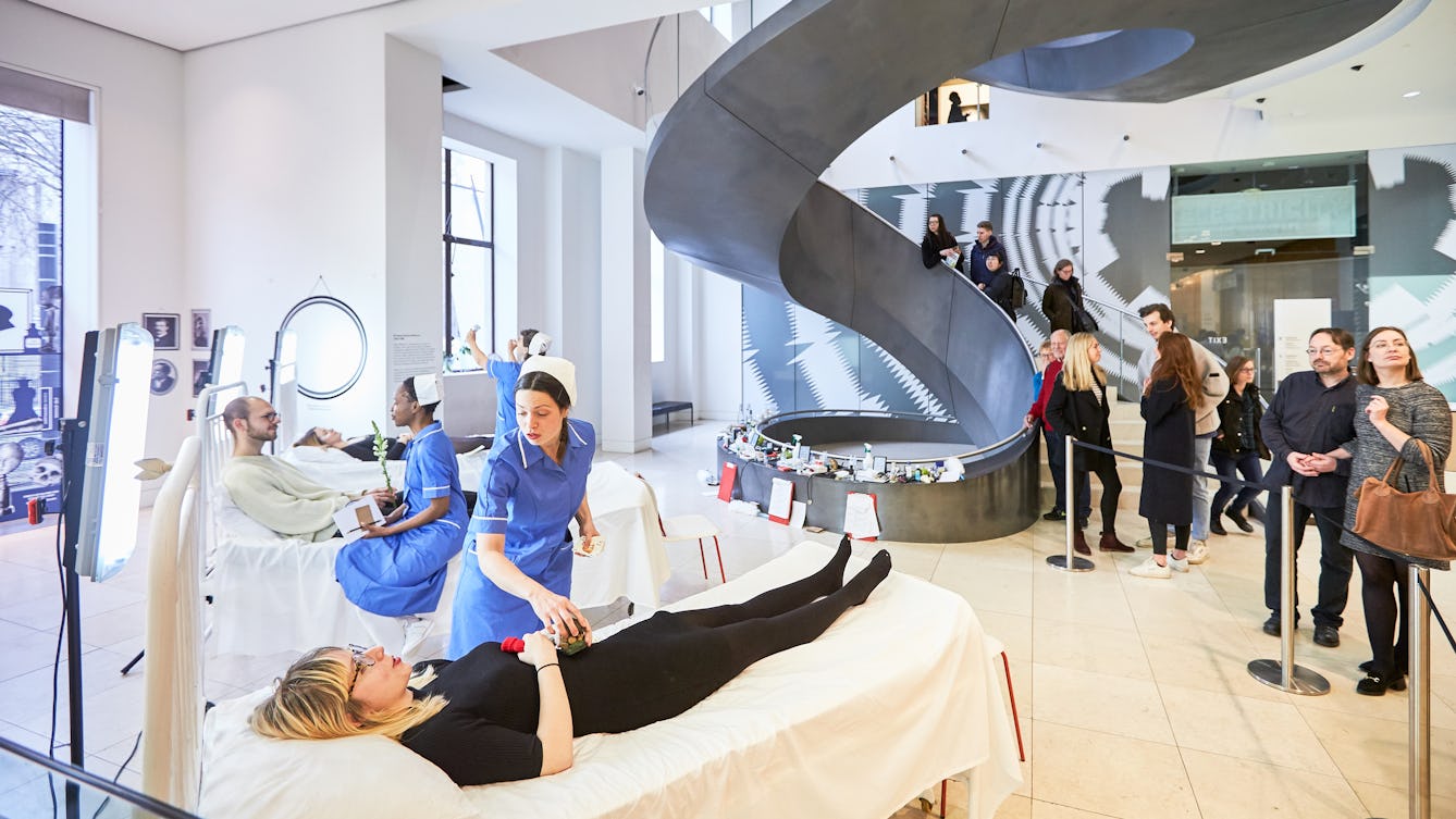 Photograph of the atrium at Wellcome Collection showing people lying on beds in the foreground attended by people wearing nurses uniforms and people watching in the background.