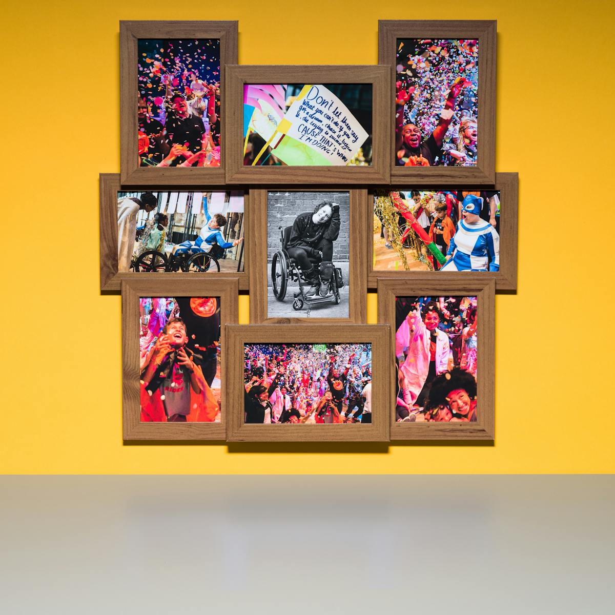 Photograph of a multiframe photo frame containing nine photographs, one in black and white and eight in colour. The frame is hung on a yellow wall above a grey tabletop.