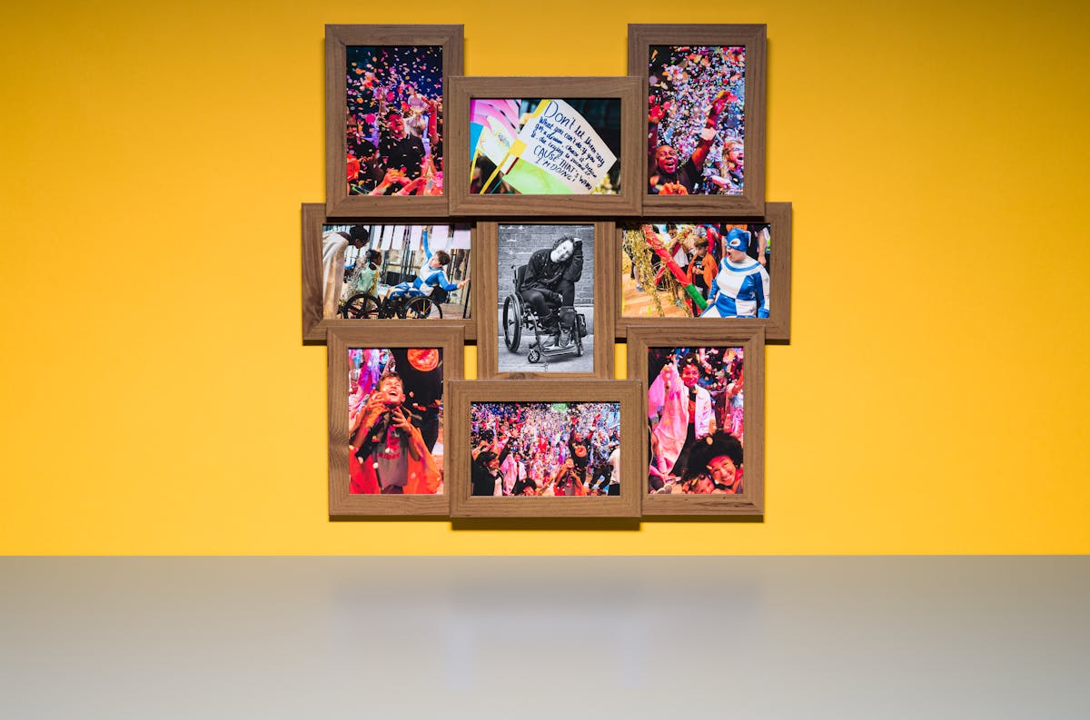 Photograph of a multi-frame photo frame containing nine photographs, one in black and white and eight in colour. The frame is hung on a yellow wall above a grey tabletop.