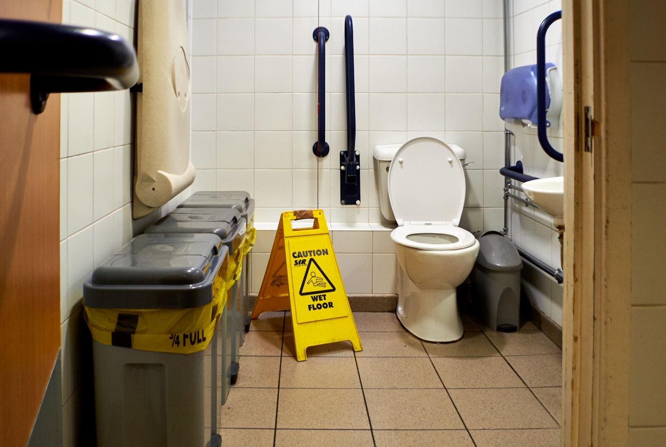 Photograph though the doorway of an accessible toilet showing part of the door and doorframe, along with the contents of the room, toilet, basin and sanitary bins. The room looks run down and dirty. To the left of the toilet is a yellow, caution wet floor sign.