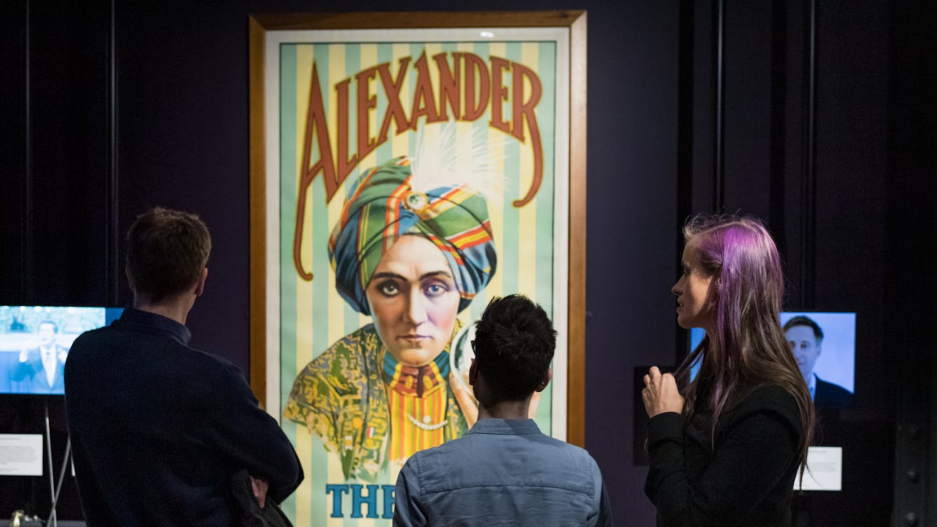 Photograph of three people looking at a large framed poster of Alexander the magician, as part of the Smoke and Mirrors exhibition at Wellcome Collection