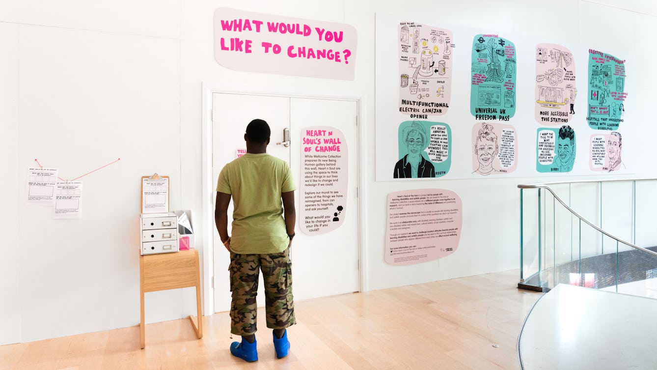 Photograph of a man looking at an exhibition installation on a gallery hoarding, made up of hand drawn text and illustration. The large pink title reads, 'What would you like to change?'. 