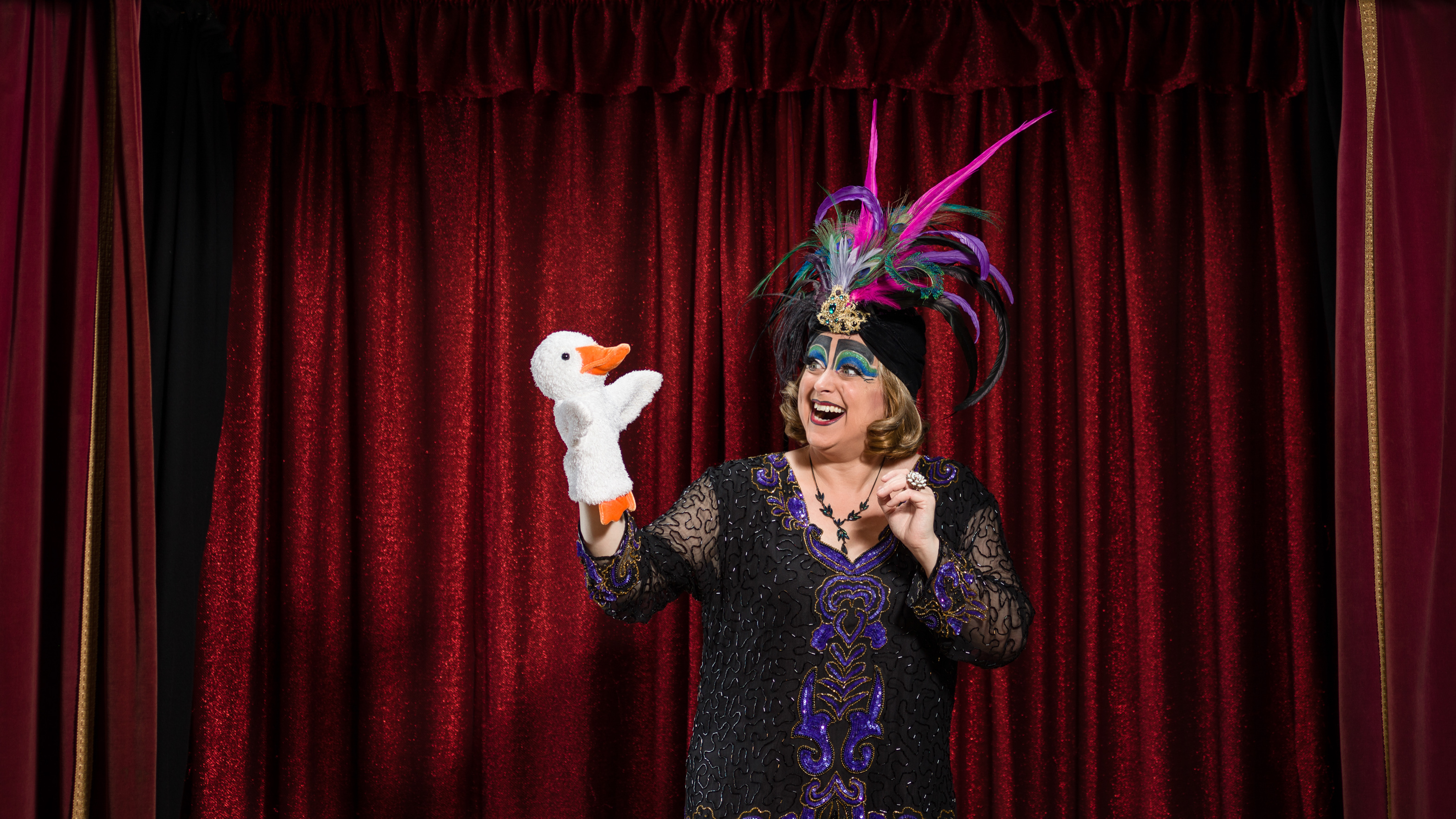 Photograph of a woman wearing a colourful feathered headdress and character makeup, looking at a white glove puppet of a duck in her hand, in front of the red velvet curtains of a theatrical stage set.