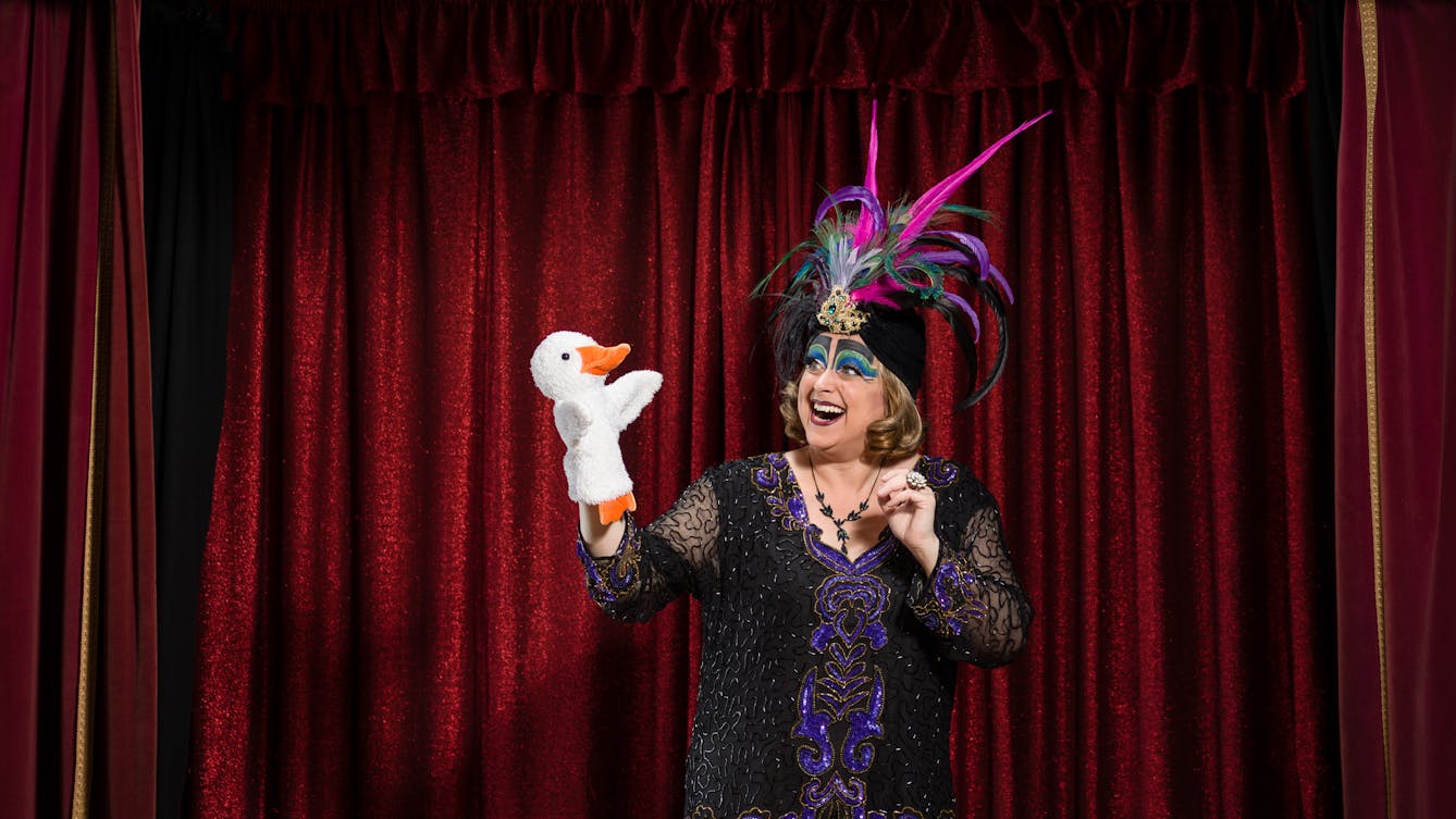 Photograph of a woman wearing a colourful feathered headdress and character makeup, looking at a white glove puppet of a duck in her hand, in front of the red velvet curtains of a theatrical stage set.
