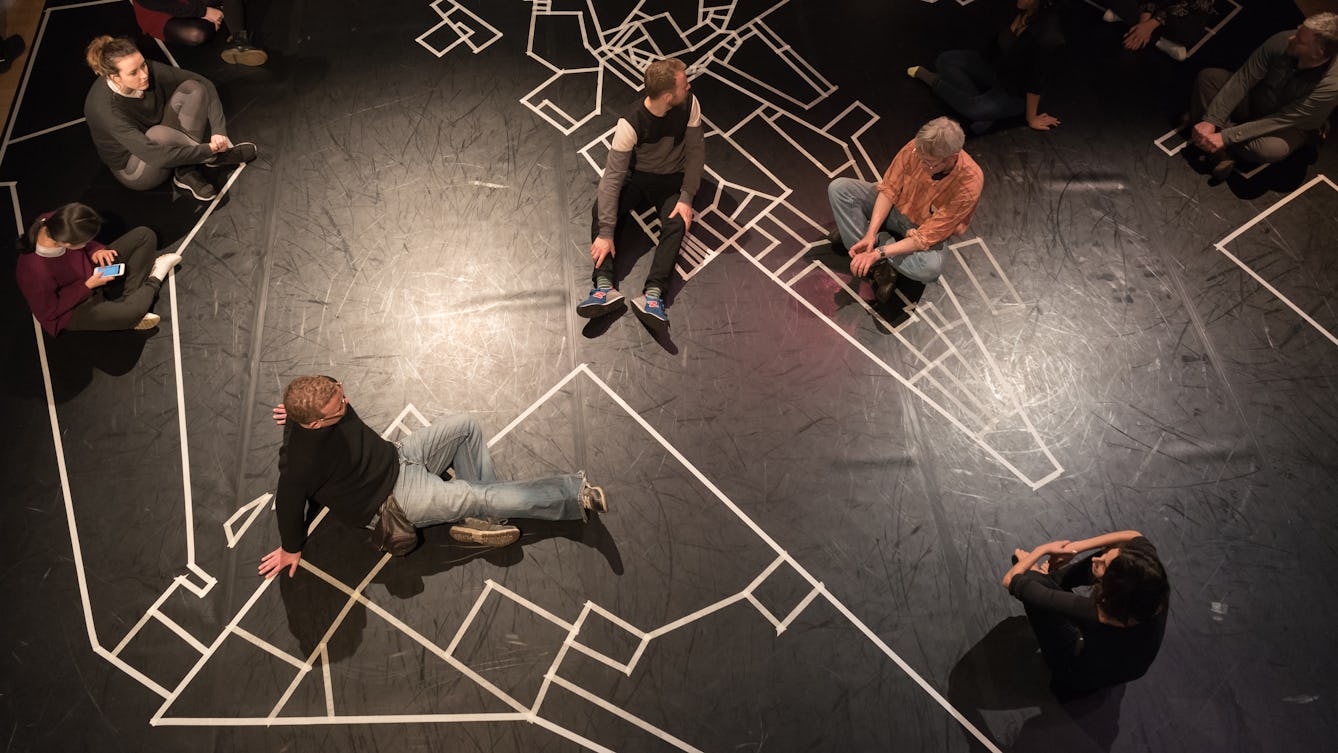 Photograph of an aerial view of a group of people sitting on the floor. The floor is black with white tape forming a giant map.