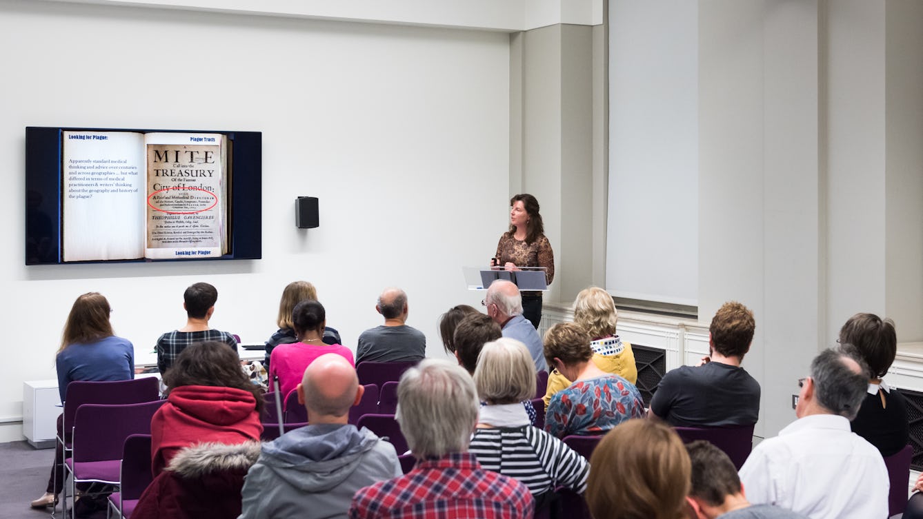 Photograph showing a woman giving a talk in the Viewing Room at Wellcome Collection. She is stood at the front of the room looking at a wall mounted television screen. In the foreground are the backs of the heads of the audience.