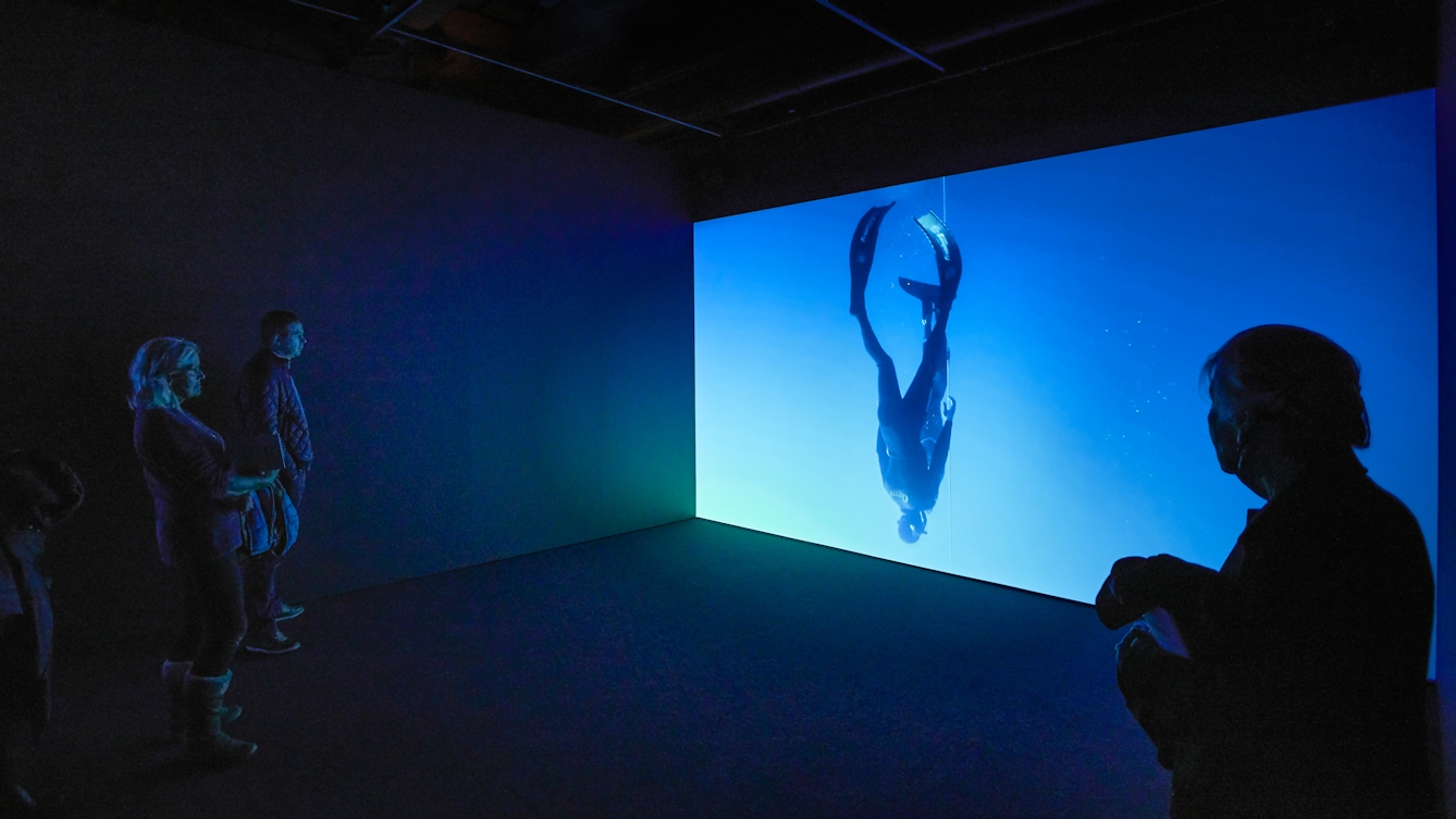Photograph of a dark gallery space with one wall lit up with a projected film of a diver. In the foreground are visitors looking at the film.