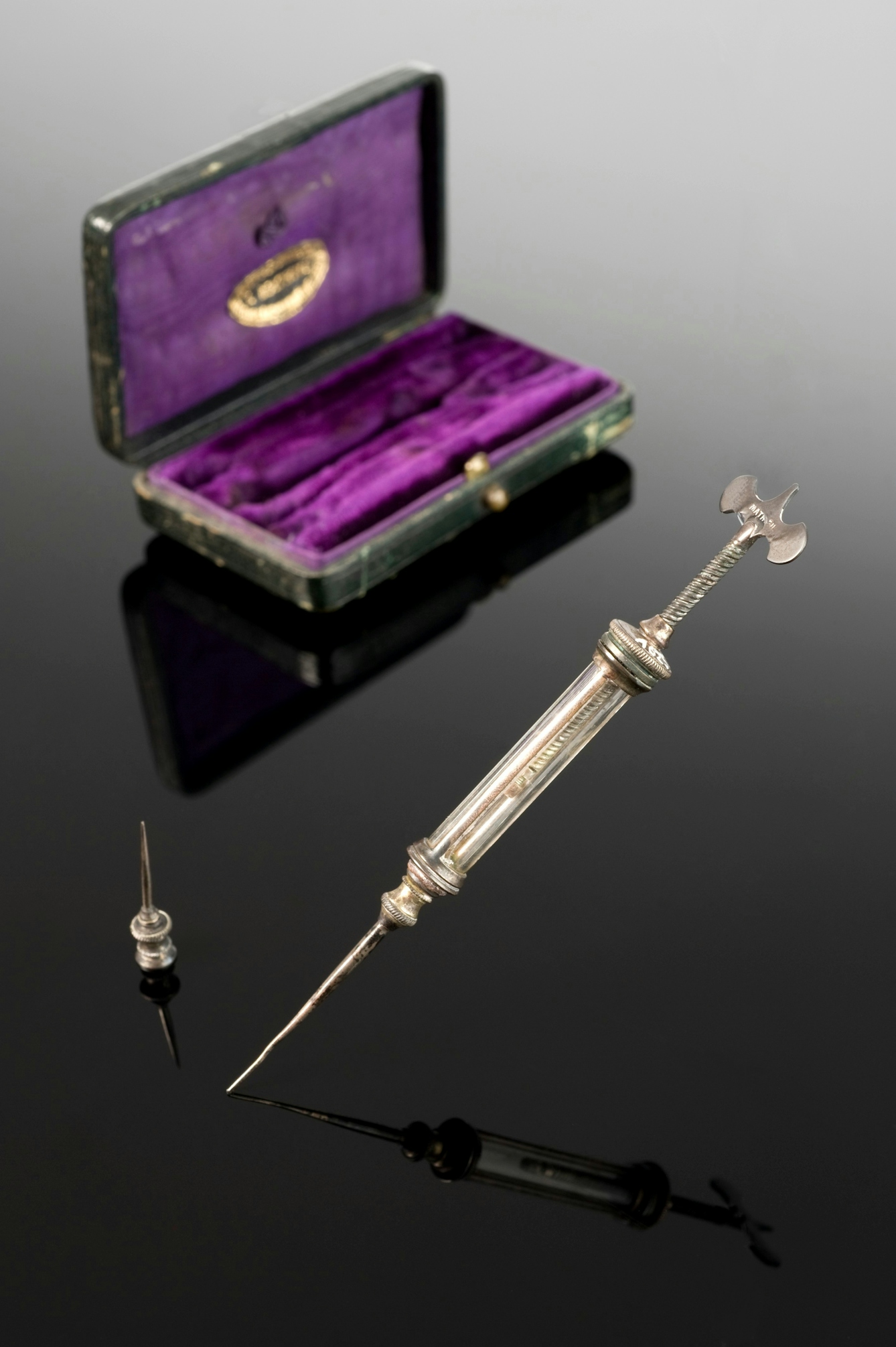 Metal syringe with a spare needle and (in background) a purple-velvet lined case.