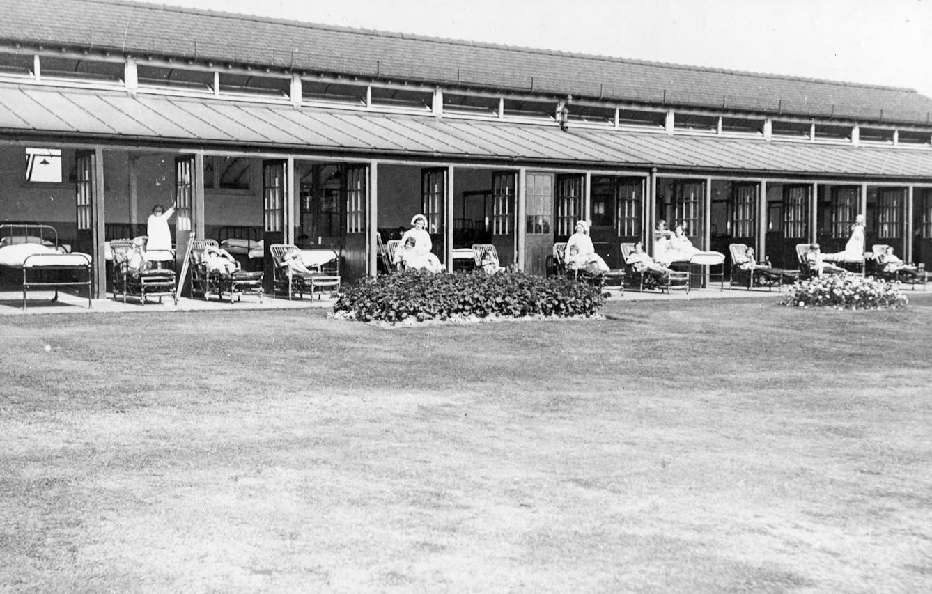 Shows patients and staff at a sanatoria in the West Midlands. Patients are sat outdoors, while accompanied by staff. 
