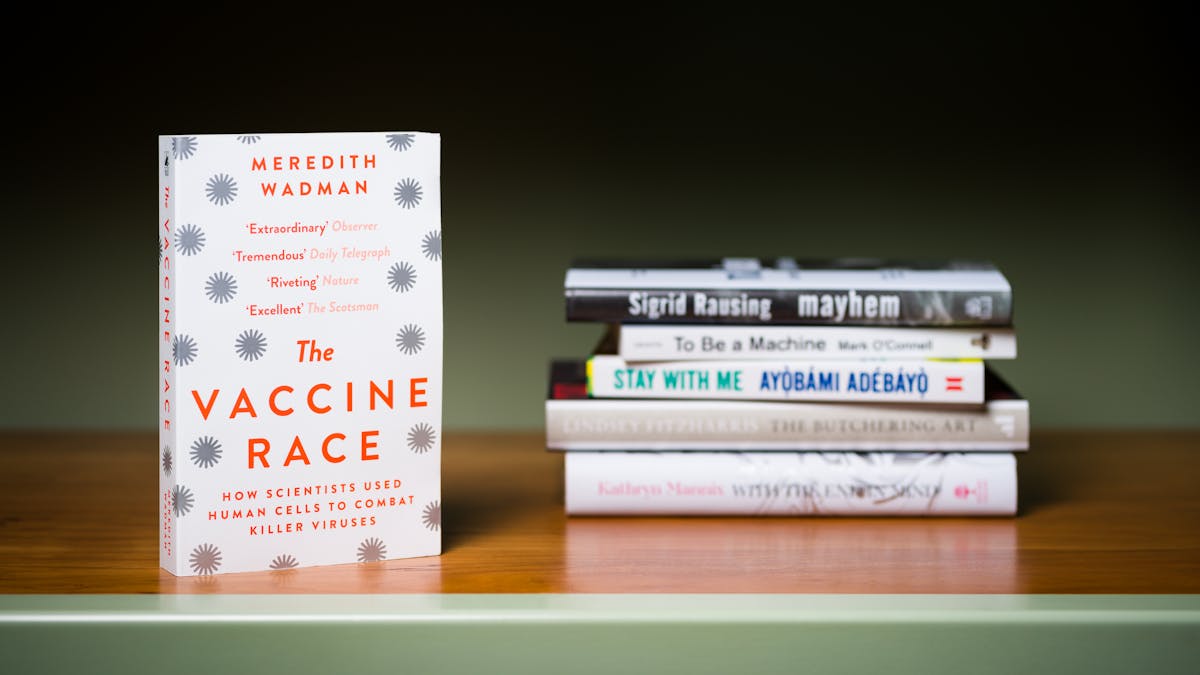 Photograph of a a stack of 5 books lying horizontally on a shelf with another book standing  vertically in the foreground.