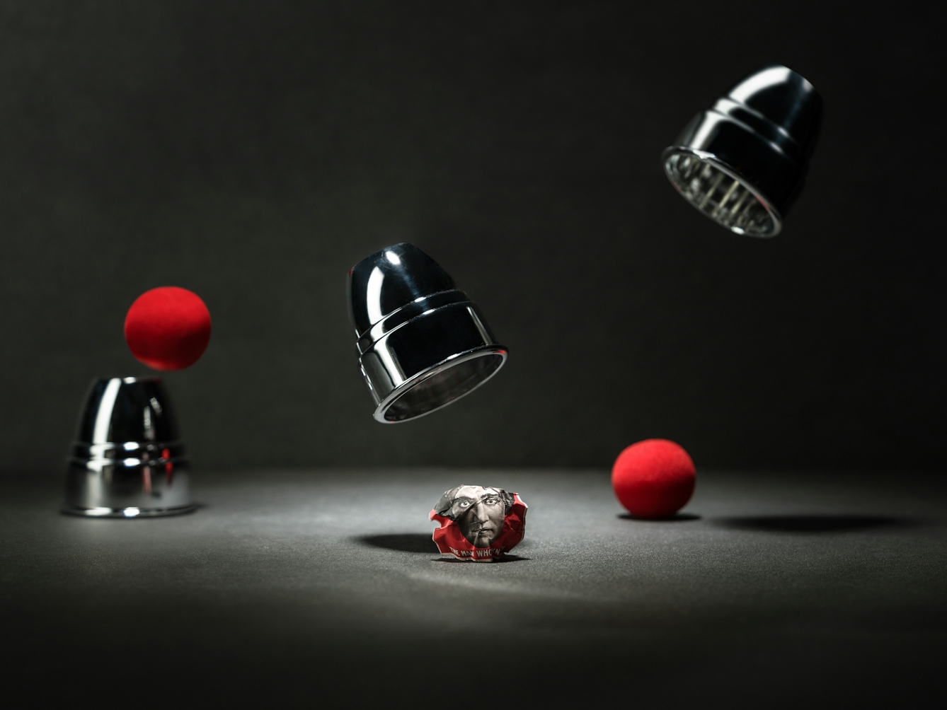 Photograph of a magic cup and ball trick with two red balls. Two of the cups are floating in mid-air. On the table is a scrunched up poster showing the face of the 1920s mind reader, Alexander.