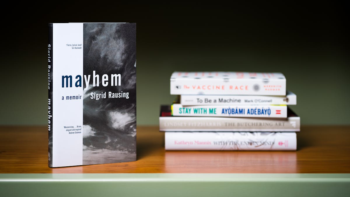 Photograph of a a stack of 5 books lying horizontally on a shelf with another book standing vertically in the foreground.