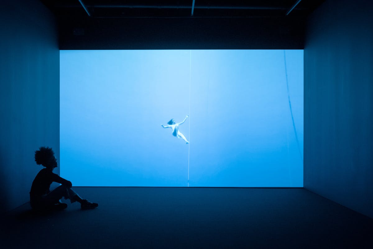 Photograph of a visitor exploring a gallery installation. She is sitting in the dark on the floor, in front of a large projection of a figure floating underwater. The room has a blue cast.
