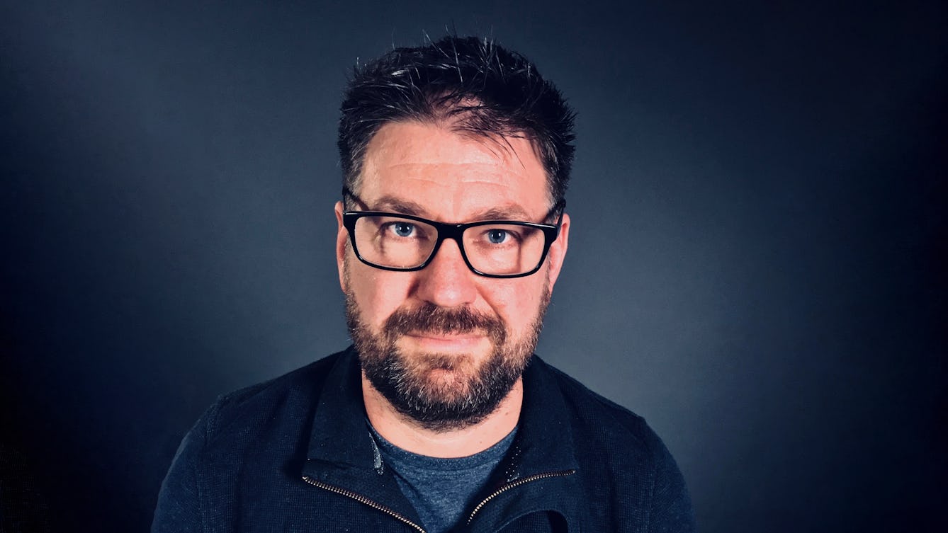 Photograph of the head and shoulders of a man with a beard and glasses against a graduated blue background.