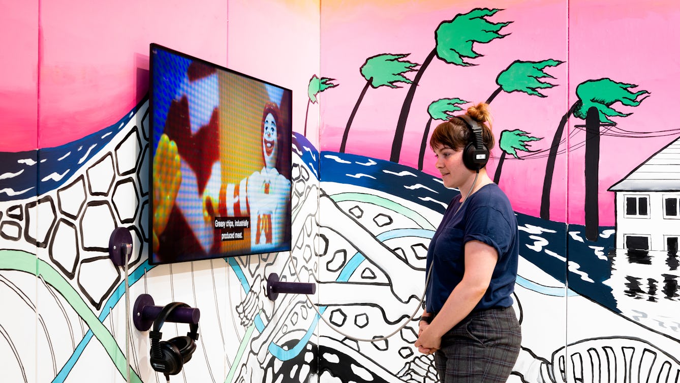 Photograph of RawMinds Art Wall in the entrance of Gallery 1 in Wellcome Collection. In the photograph a woman is wearing headphones and watching a film. In the background is a colourful installation, inspired by climate change and the ‘Being Human‘.