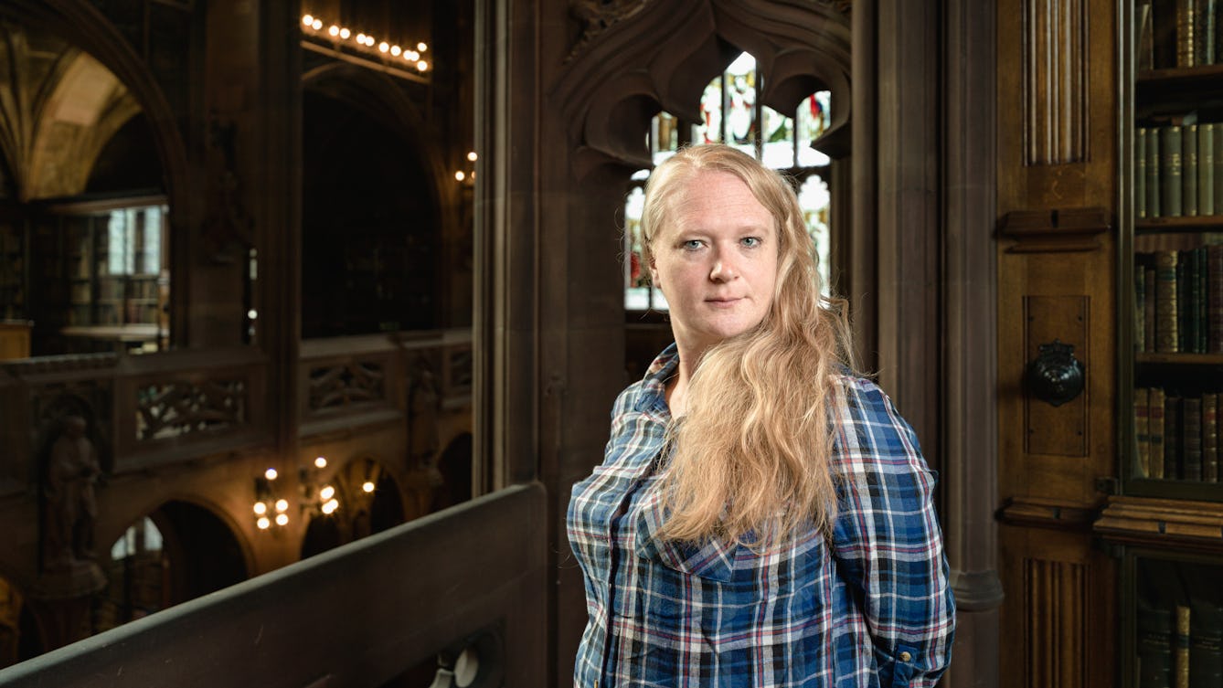 Photograph of Dr Joanne Edge standing in the John Rylands Library in Manchester.  In the background, there are wooden shelves with books and a stained glass window.