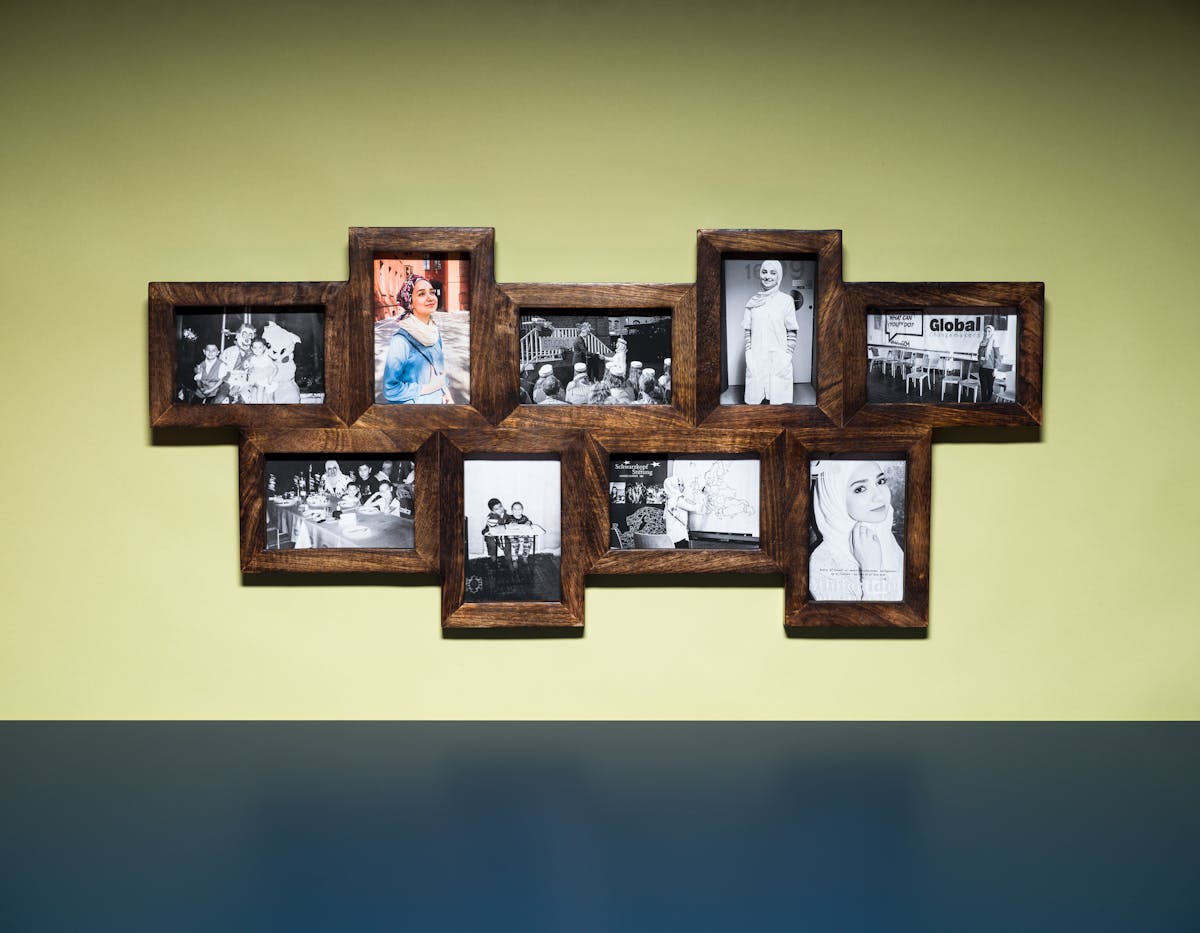 Photograph of a multi-frame photo frame containing nine photographs, eight in colour and one in black and white. The frame is hung on a light green coloured wall above a blue tabletop.