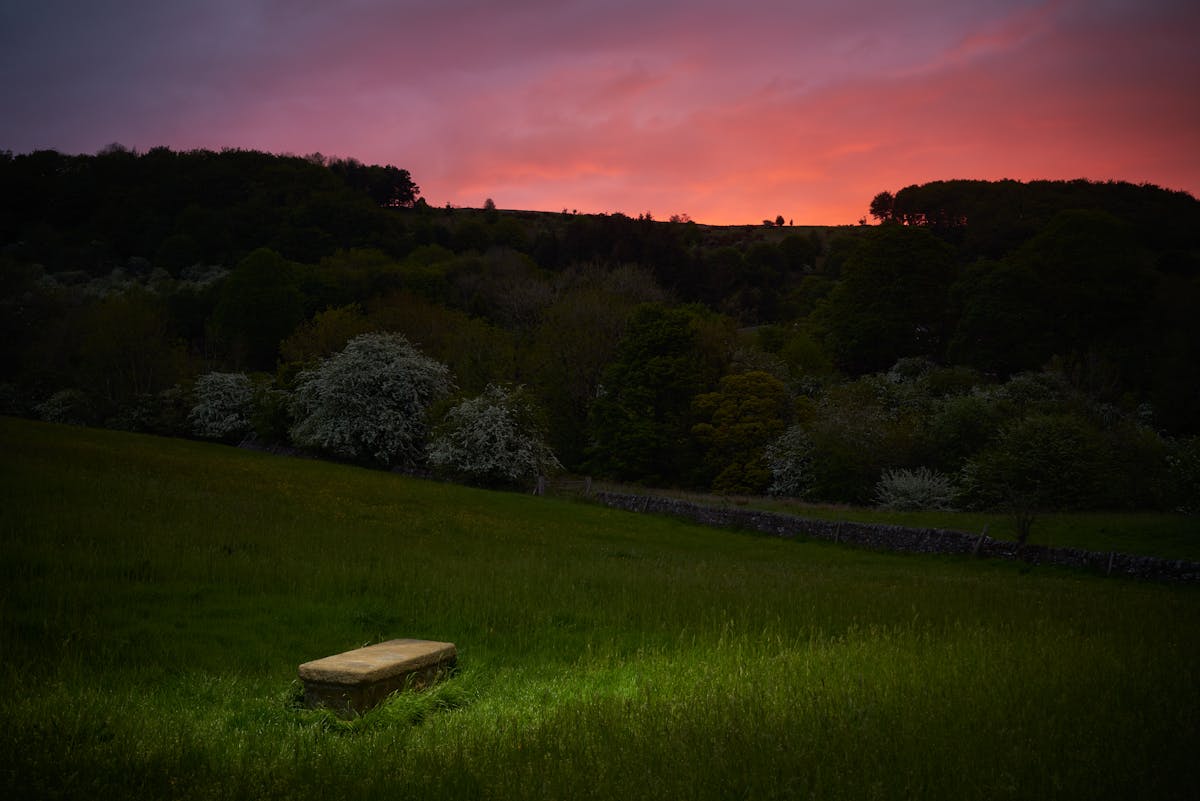 Photograph of a rural landscape at dusk. In the background is a dramatic red sky. In the foreground is a spotlit plain stone grave.