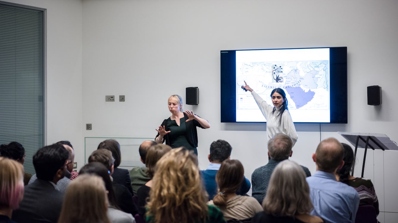 Photograph of Aarathi Prasad pointing at a large television screen in the Viewing Room at Wellcome Collection during a Library Insights event. There is a British sign language interpreter to the left facing the audience. The backs of the audience's heads are in the foreground.