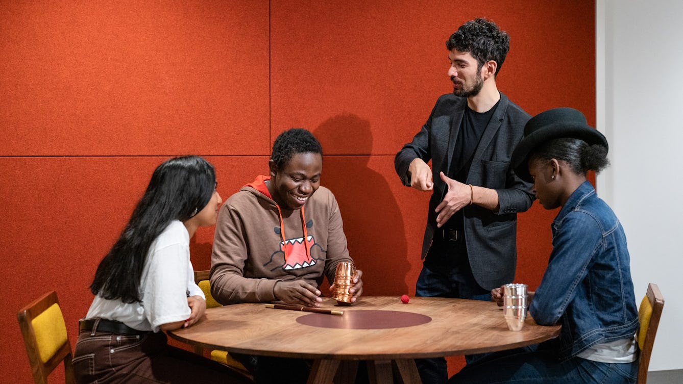 Photograph of 3 young participants sat around a table. One participant is being taught a magic trick by a magician standing next to them. On the table are magic tick cups and balls.