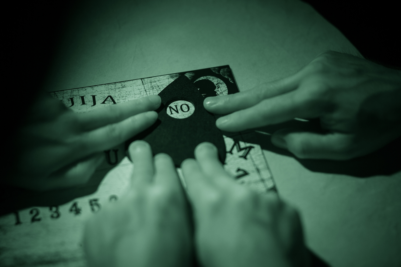 Photograph of a ouija board with the hands of three people resting on the planchette. The image is toned green as a result of being made under infrared light.