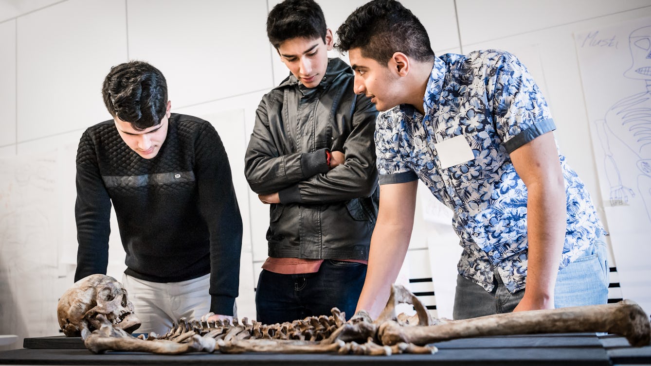 Photograph of three young men discussing a skeleton which is laid out on a table in front of them.