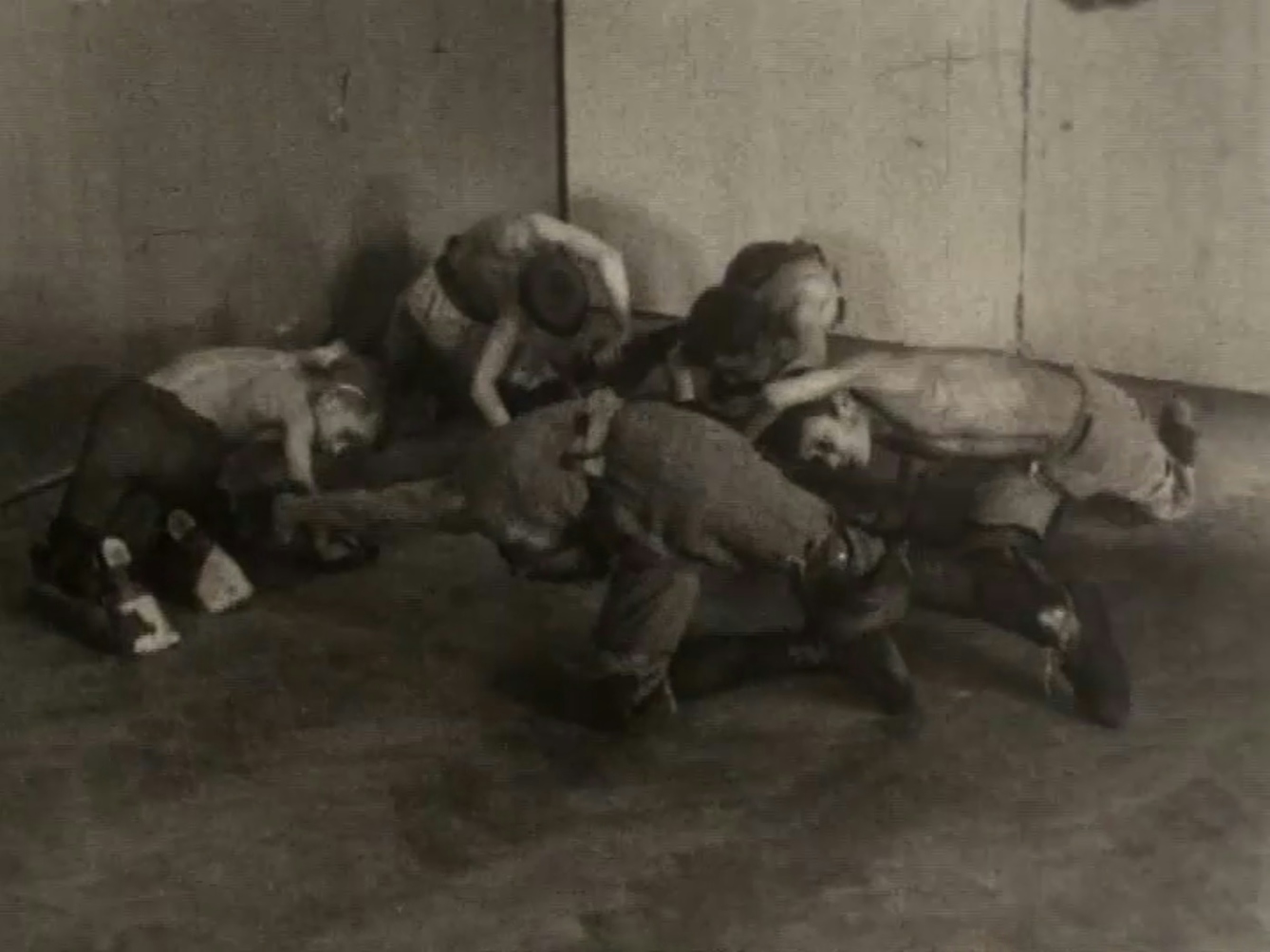 Black and white still from a film showing boys crawling around in a circle on the floor, arching their arms over their heads as if swimming.