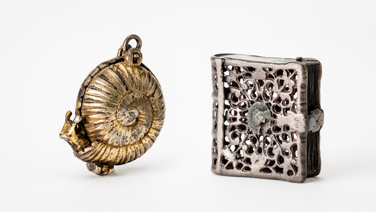 Two pomanders photographed against a white background; one is gold coloured and snail shaped, the other is silver coloured and book shaped.