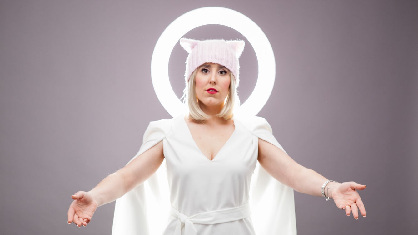 Photograph of Lee Minora from the waist up, wearing a white dress with white cape. She is wearing a wooly hat with ears and is holding our her arms to the side. Behind her head is a halo.