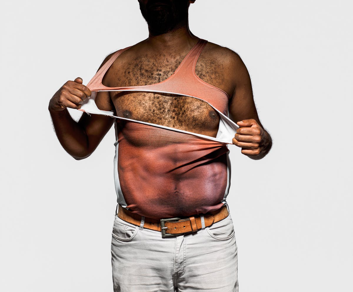 Photograph of a man from the thigh up wearing a vest which has a very muscly male torso printed on it. The man is ripping the vest open to reveal his own torso beneath. His face is hidden in shadow.