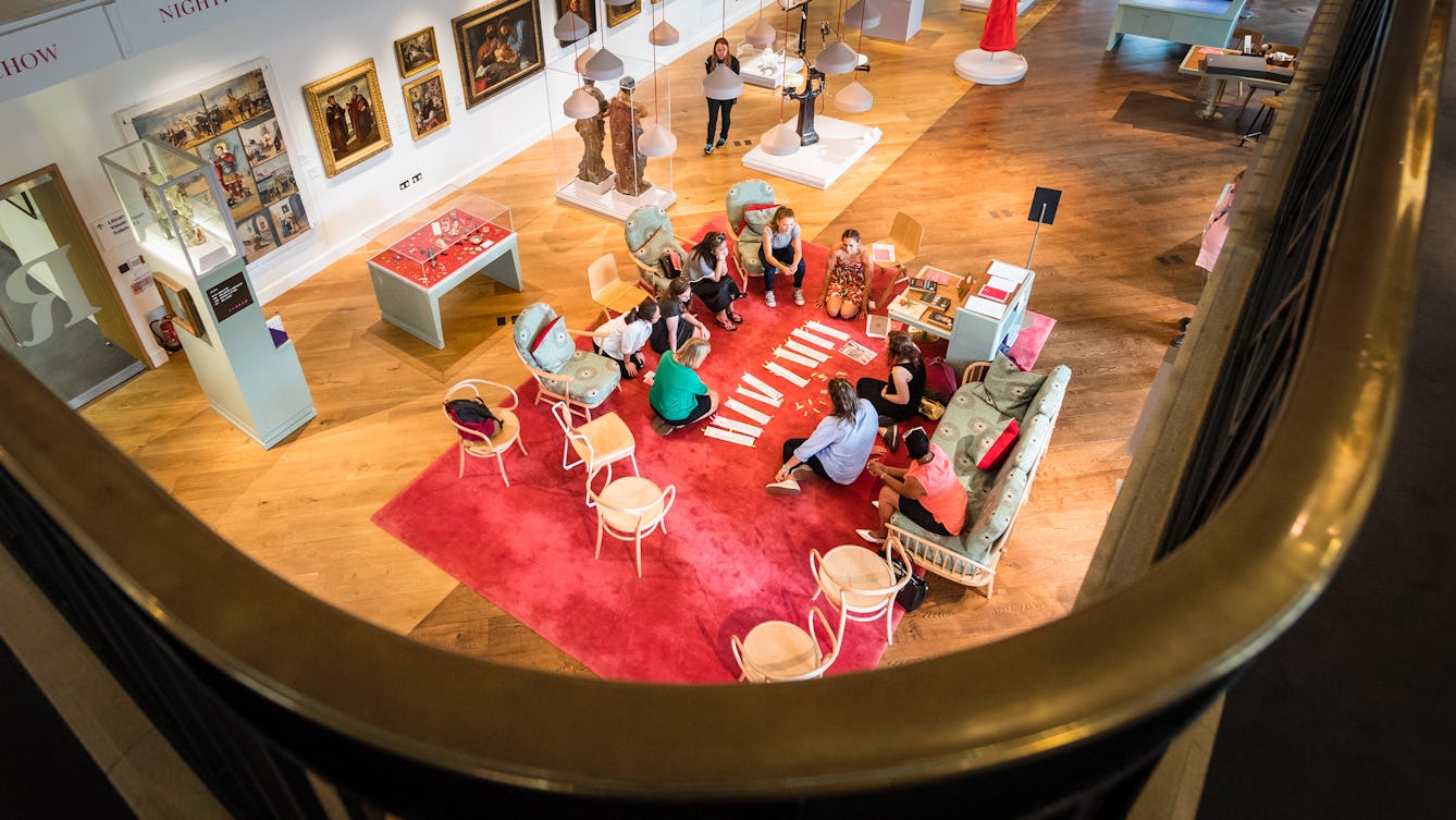 Photograph from the gallery level in the Reading Room looking down into the exhibition space below. There is a group of eight people sitting on a carpet engaged in an activity involving strips of paper containing typed words.